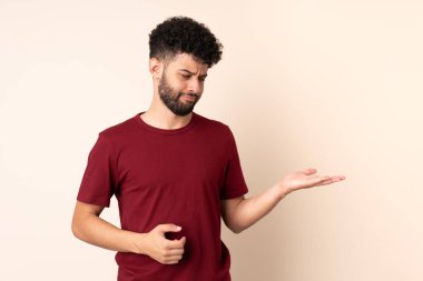 Faslı genç bir adam bej arka planda izole edilmiş. Kopyalama alanını şüphe ile tutuyor.