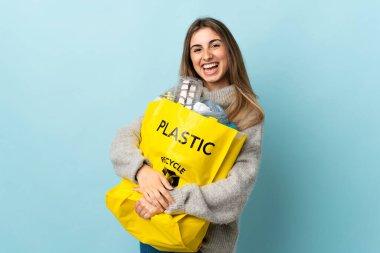 Geri dönüşüm için plastik şişelerle dolu bir torba tutarak yalıtılmış maviliğin üzerinde yan tarafa bakıyor.