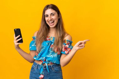 İzole edilmiş sarı arka planda cep telefonu kullanan genç bir kadın şaşırmış ve işaret ediyor.
