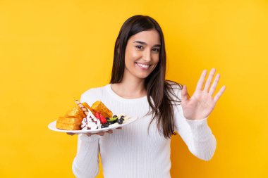 Genç esmer kadın, izole edilmiş arka planda waffle tutuyor mutlu bir ifadeyle selamlıyor.