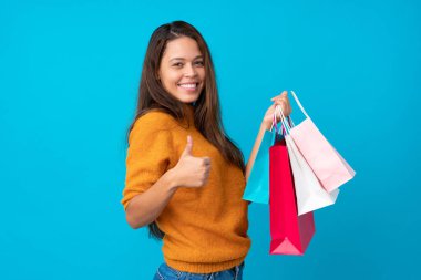 İzole edilmiş mavi arka planda genç Brezilyalı kız elinde alışveriş çantaları ve başparmağıyla