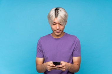 Asyalı genç adam izole edilmiş mavi arka planda cep telefonuyla mesaj gönderiyor.