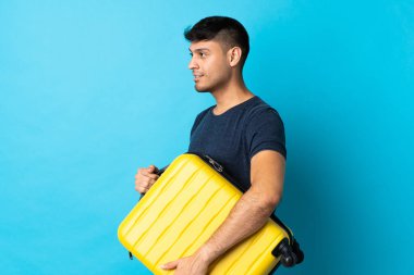 Genç Kolombiyalı, mavi arka planda izole edilmiş seyahat valiziyle tatilde.