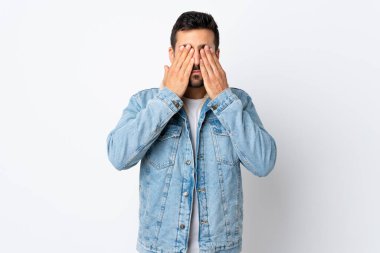 Beyaz arka planda sakallı, yakışıklı genç bir adam. Gözlerini elleriyle kapatıyor.