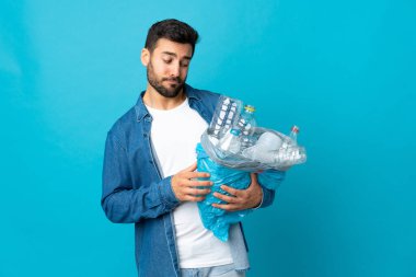 Mavi arka planda izole edilmiş bir şekilde geri dönüşüm için plastik şişe dolu bir çanta taşıyan genç beyaz bir adam.