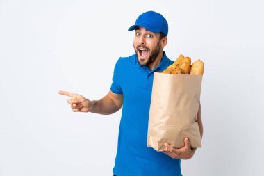 Elinde bir çanta dolusu ekmek tutan teslimatçı beyaz arka planda izole edilmiş şaşırmış ve parmağını kenarı işaret ediyor.