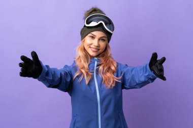 Kar kayağı gözlüklü genç kız izole edilmiş mor arka planda sunuyor ve davet ediyor.