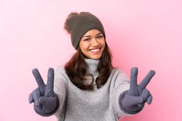 Young Colombian girl with winter hat over isolated pink wall smiling and showing victory sign