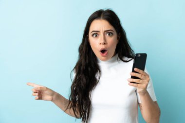 Mavi arka planda izole edilmiş cep telefonu kullanan genç bir kadın şaşırmış ve işaret ediyor.