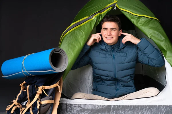 Teenager caucasian man inside a camping green tent isolated on black background frustrated and covering ears