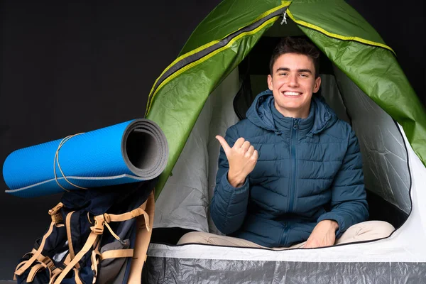 Teenager caucasian man inside a camping green tent isolated on black background pointing to the side to present a product
