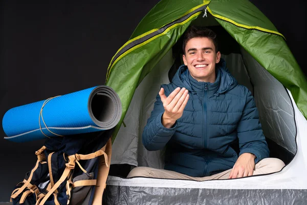 Teenager caucasian man inside a camping green tent isolated on black background inviting to come with hand. Happy that you came