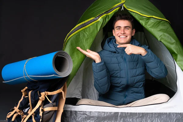 Teenager caucasian man inside a camping green tent isolated on black background holding copyspace imaginary on the palm to insert an ad