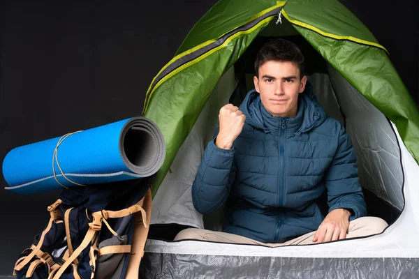 Teenager caucasian man inside a camping green tent isolated on black background with angry gesture