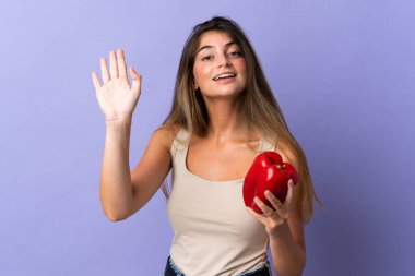 Mor arkaplanda biber tutan genç bir kadın. Mutlu bir ifadeyle selamlıyor.