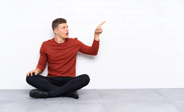 Mladý Pohledný Muž Sedí Podlaze Ukazuje Pryč — Stock fotografie