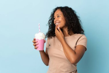 Çilekli milkshake 'i olan genç kız mavi arka planda izole edilmiş, yan tarafa bakıyor ve gülümsüyor.