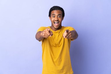 Örgülü genç Afrikalı Amerikalı adam mor arka planda izole edilmiş şaşırmış ve ön tarafı işaret ediyor.