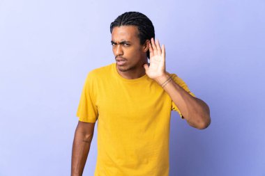 Örgülü genç Afrikalı Amerikalı adam mor arka planda izole edilmiş. Kulağına elini sürerek bir şey dinliyor.