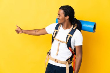 Sarı arka planda izole edilmiş büyük bir sırt çantalı genç dağcı, baş parmak hareketi yapıyor.