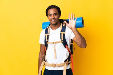 Sarı arka planda izole edilmiş büyük bir sırt çantalı genç dağcı mutlu bir ifadeyle selamlıyor.