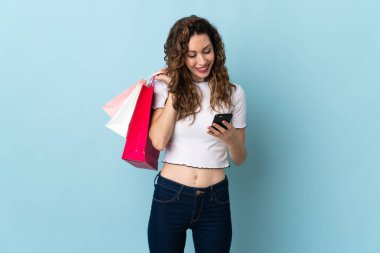 Kafkasyalı genç bir kadın mavi arka planda izole edilmiş. Elinde alışveriş poşetleri var ve bir arkadaşına cep telefonuyla bir mesaj yazıyor.