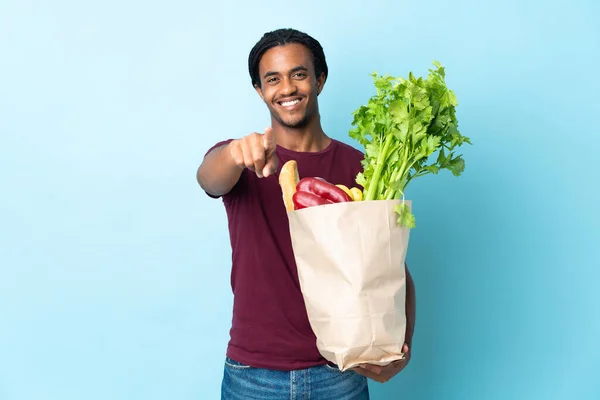 African American man holding a grocery shopping bag isolated on blue background pointing front with happy expression