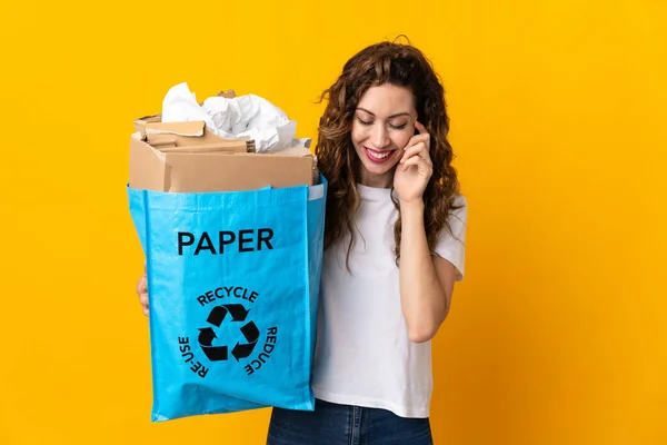 リサイクルバッグを持っている若い女性は 黄色の背景笑いに隔離されたリサイクルするために紙でいっぱい — ストック写真