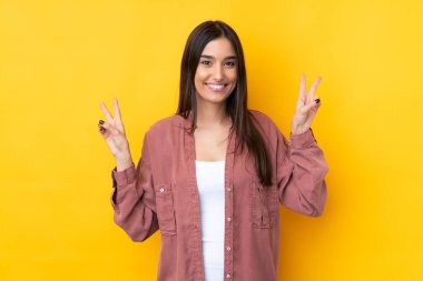 Genç esmer kadın izole edilmiş sarı arka planda iki eliyle zafer işareti gösteriyor.