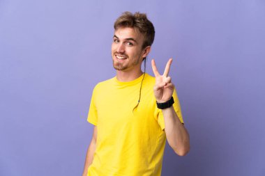 Mor arka planda izole edilmiş yakışıklı sarışın adam gülümsüyor ve zafer işareti gösteriyor.