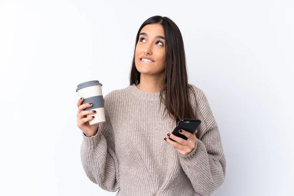 年轻的黑发女人在孤独的白色背景上一边拿着咖啡一边想着什么 一边拿着手机 — 图库照片