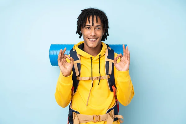若いです登山家アフリカ系アメリカ人男性とともに大きなバックパックで隔離された青い背景に指でOk記号を示す — ストック写真