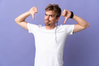 Mor arka planda soyutlanmış yakışıklı sarışın adam baş parmağını iki eliyle aşağı gösteriyor.