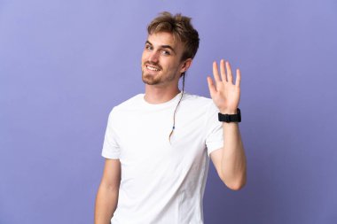 Mor arka planda izole edilmiş genç, yakışıklı sarışın adam mutlu bir ifadeyle selamlıyor.