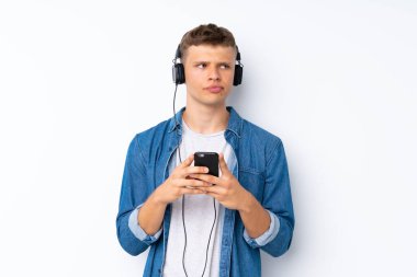 Soyutlanmış beyaz arka planda genç yakışıklı adam kulaklıklı ve düşünceli cep telefonunu kullanıyor.