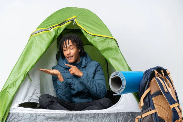 Young african american man inside a camping green tent holding copyspace imaginary on the palm to insert an ad