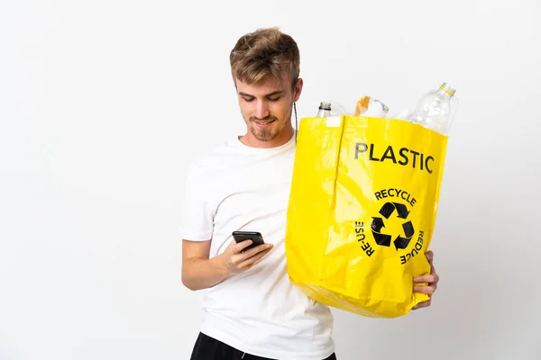 Young blonde man holding a recycling bag full of paper to recycle isolated on white background sending a message with the mobile