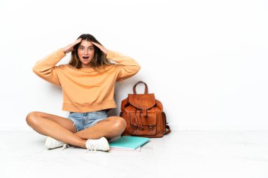 Yerde dizüstü bilgisayarlı genç bir bayan oturuyor. Beyaz arka planda, sürpriz bir ifadeyle izole edilmiş.