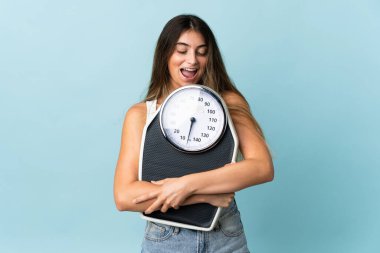 Genç beyaz kadın, mavi arka planda, tartı makinesiyle izole edilmiş.