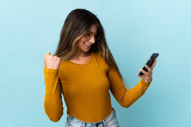 Mavi arka planda izole edilmiş cep telefonu kullanan beyaz bir kadın zaferi kutluyor.