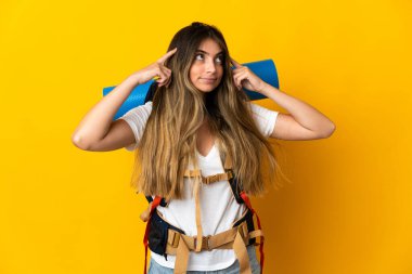 Sarı arka planda izole edilmiş büyük sırt çantalı genç dağcı kadın kuşkuları ve düşünceleri var.