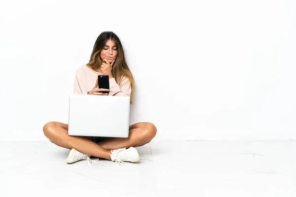 Young woman with a laptop sitting on the floor isolated on white background thinking and sending a message