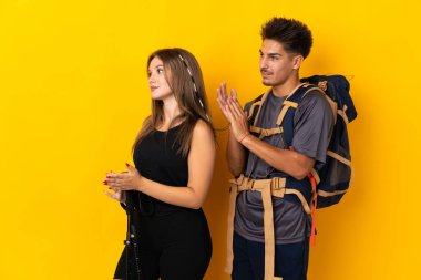 Sarı arka plana izole edilmiş büyük sırt çantalı genç dağcı çift bir konferansta sunumdan sonra alkışlıyorlar.