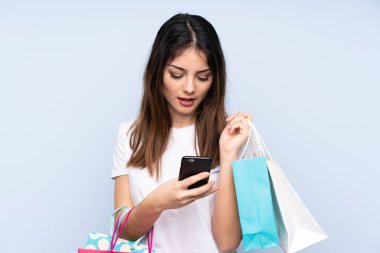 Genç esmer kadın izole edilmiş mavi arka planda elinde alışveriş torbalarıyla bir arkadaşına cep telefonuyla bir mesaj yazıyor.