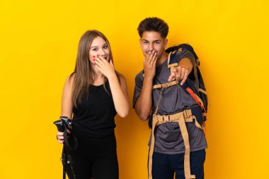 Sarı arka planda izole edilmiş büyük sırt çantalı genç dağcı çifti birine işaret ediyor ve gülüyor.
