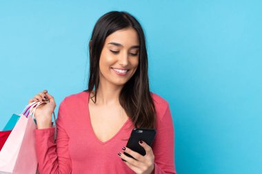 Genç esmer kadın izole edilmiş mavi arka planda elinde alışveriş torbalarıyla bir arkadaşına cep telefonuyla bir mesaj yazıyor.