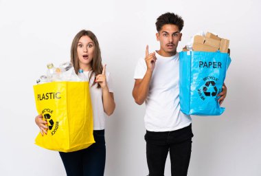 Beyaz arka planda izole edilmiş plastik ve kağıt dolu bir çanta tutan genç çift, çözümü fark etmek için parmağını kaldırıyor.