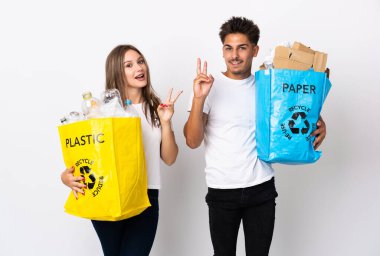 Beyaz arka planda izole edilmiş plastik ve kağıt dolu bir çanta taşıyan genç çift gülümsüyor ve zafer işareti gösteriyor.
