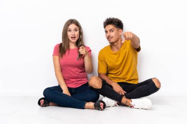 Yerde oturan genç çift beyaz arka planda izole edilmiş şaşırmış ve ön tarafı işaret ediyor.