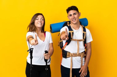 Sarı arka planda izole edilmiş büyük sırt çantalı genç dağcı çift parmağını kaldırıyor.